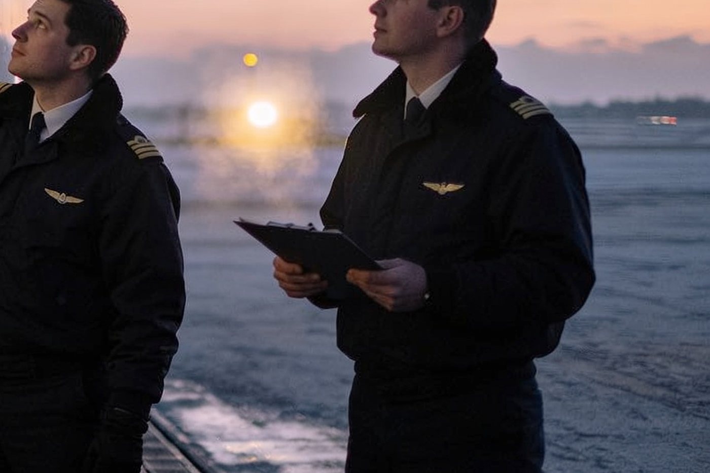 Two pilots in dark uniform jackets standing on a frosted ramp at first light, one holding a clipboard, both looking up at the aircraft above them.