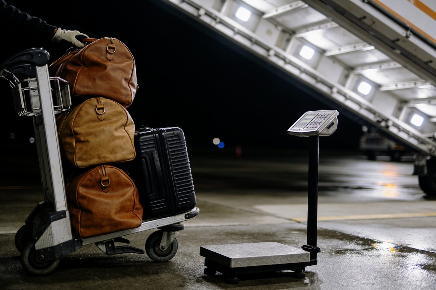Three leather duffel bags and a hard case stacked on a baggage cart beside a platform scale on a wet ramp at night, a gloved hand steadying the top bag, airstairs behind.